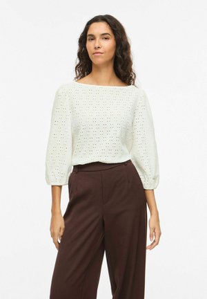 Kobieta o długich kręconych włosach, ubrana w białą bluzkę z pufiastymi rękawami i wysokim stanem, ciemnobrązowe spodnie z szerokimi nogawkami, stojąca.