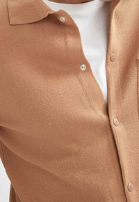 Gros plan d'une personne portant un cardigan en tricot beige avec des boutons-pression sur un tee-shirt blanc à col rond.