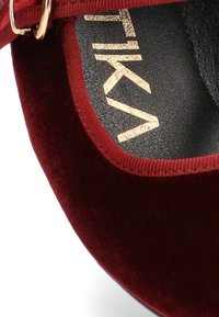 Zapato de terciopelo burdeos con una plantilla negra texturizada. Presenta una correa estrecha y detalles en oro del logo. Textura suave con un diseño de puntera redondeada.