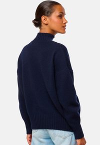 Femme avec les cheveux en chignon portant un pull à col montant bleu marine et un jean bleu clair, de profil contre un fond uni.