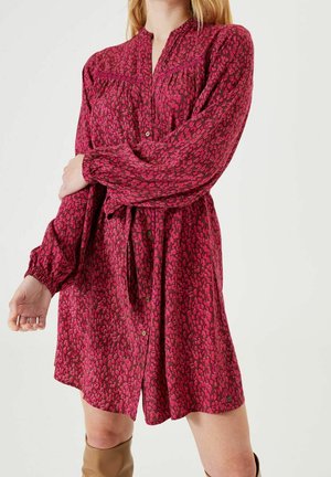 Femme portant une robe à manches longues, boutonnée sur le devant, de couleur rose et rouge foncé avec un motif, et une ceinture à la taille, debout devant un fond clair uni.