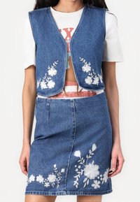 Denimset bestående av väst och kjol med blommigt vitt broderi, ärmlös design och en figurnära silhuett. Ljusblå denim med en mjuk textur.