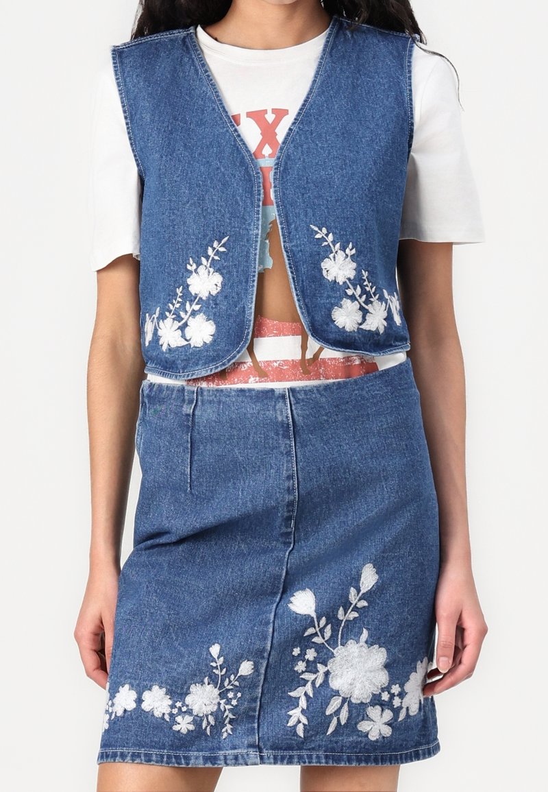 Denimset bestående av väst och kjol med blommigt vitt broderi, ärmlös design och en figurnära silhuett. Ljusblå denim med en mjuk textur.