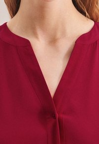 Bordová blůza s výstřihem do V, detailním zapínáním na knoflíky a hladkou strukturou. Vyznačuje se mírným leskem a čistým, minimalistickým designem.