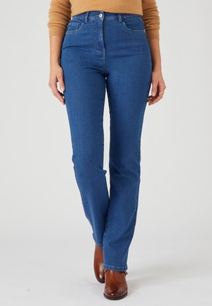 Femme portant un jean bleu taille haute et droit, des bottines marron et un haut à manches longues beige, debout devant un fond uni.