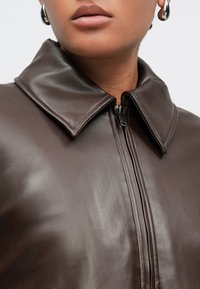 Καφέ δερμάτινο μπουφάν με μεγάλο γιακά και φερμουάρ μπροστά. Η υφή είναι λεία, υπογραμμίζοντας έναν εφαρμοστό σχεδιασμό. Τα στοιχεία περιλαμβάνουν κλείσιμο με φερμουάρ.