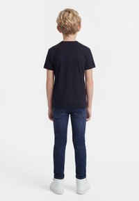 Niño rubio de pie de espaldas, vestido con una camiseta negra de manga corta, jeans azul oscuro y zapatillas blancas sobre un fondo blanco.