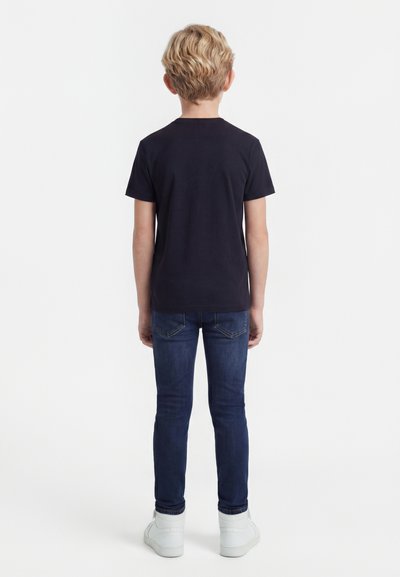 Ξανθός αγόρι που στέκεται γυρισμένο προς τα πίσω, φοράει μαύρο κοντομάνικο πουκάμισο, σκούρο μπλε τζιν και λευκά αθλητικά παπούτσια σε λευκό φόντο.