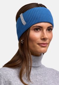 Donna con lunghi capelli castani che indossa una fascia blu a maglia con l'etichetta "FALKE", orecchini a cerchio in argento e un maglione a collo alto grigio a coste.