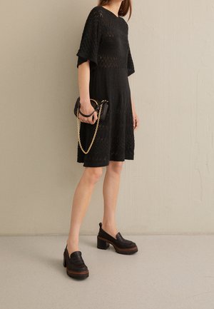 Mujer con un vestido negro de punto hasta la rodilla y mocasines negros de tacón grueso, sosteniendo un pequeño bolso negro con cadena dorada.