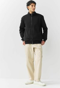 Mann in schwarzer Kapuzenjacke, beigen Hosen und weißen Sneakers, der vor einem schlichten weißen Hintergrund steht.