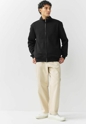 Hombre vestido con una chaqueta negra con cremallera, pantalones beige y zapatillas blancas, de pie contra un fondo blanco liso.