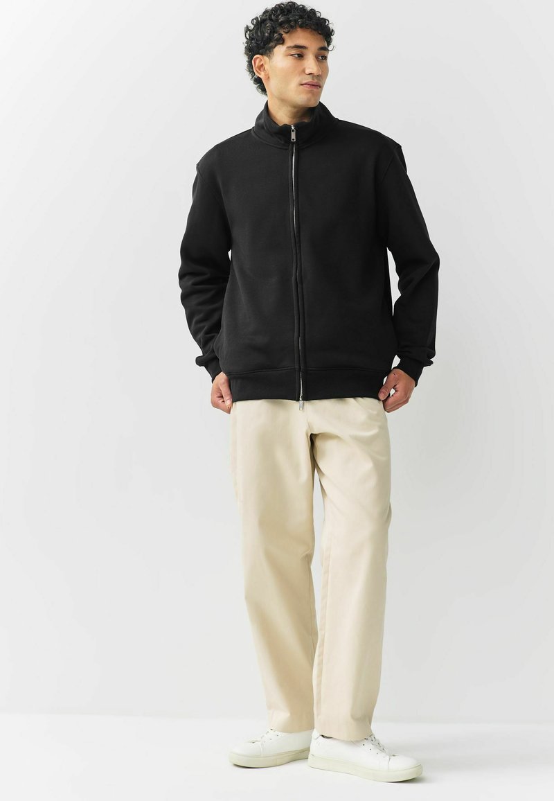 Mann in schwarzer Kapuzenjacke, beigen Hosen und weißen Sneakers, der vor einem schlichten weißen Hintergrund steht.