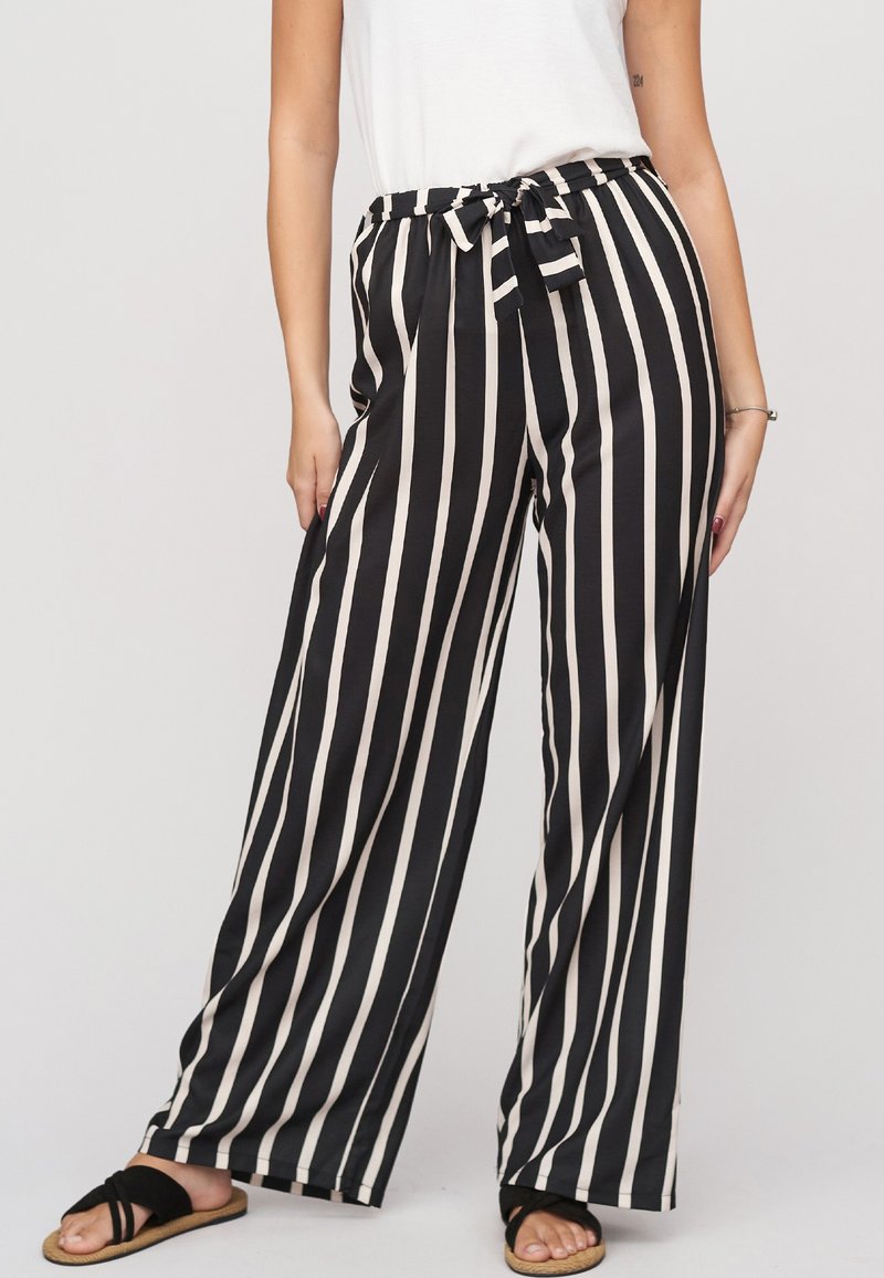 Femme portant un pantalon large à rayures verticales noires et blanches avec une taille nouée, un haut blanc et des sandales noires.