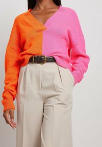 Pull en tricot avec un design fendu aux couleurs orange et rose, col en V, manches longues, associé à un pantalon plissé beige et une ceinture marron.