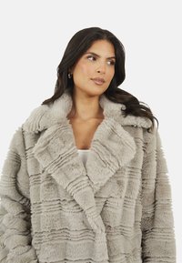 Sztuczny futrzany płaszcz w jasnoszarym kolorze z teksturowanym, prążkowanym wzorem. Posiada szeroki kołnierz i luźny krój, idealny do noszenia na warstwy.