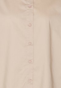 Camicia bottone color beige chiaro con una texture liscia, sei bottoni rosa coordinati sul davanti e un classico design del collo.