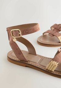 Sandales plates en daim marron avec des brides à la cheville, des boucles dorées et des détails tissés à l'avant sur un fond clair.