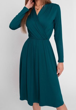 Žena nosící tyrkysové zavinovací šaty s dlouhými rukávy délky ke kolenům, se staženým pasem a zlatými vrstvenými náhrdelníky.