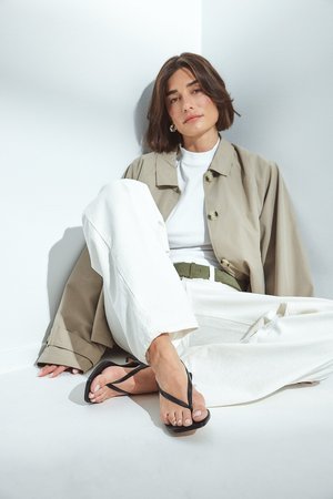 Des tongs noires avec des lanières fines, une semelle lisse, portées avec un pantalon blanc à jambes larges et une veste beige claire, sur un fond blanc uni.