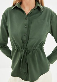 Vrouw in een donkergroene blouse met knopen, voorzien van een borstzak en een strikceintuur, gecombineerd met lichtgekleurde broek.