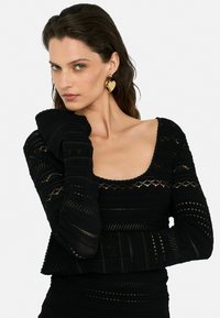Black knit top featuring textured patterns and a scoop neckline. Model wears gold heart-shaped earrings, showcasing bold design and color contrast.
