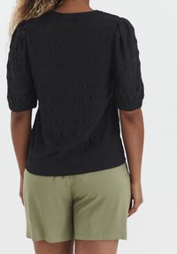 Kvinna i en texturerad svart kortärmad blus och ljus olivgröna shorts, sedd bakifrån mot en enfärgad bakgrund.