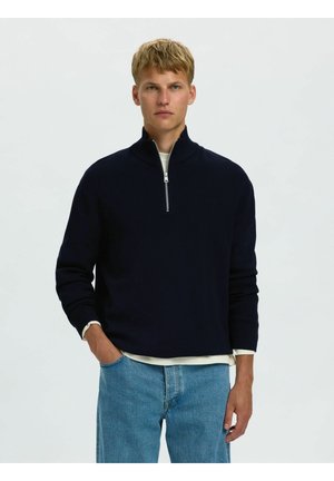 Jeune homme aux cheveux blonds courts portant un pull noir à demi-fermeture éclair par-dessus une chemise blanche et un jean bleu, debout avec une main dans la poche.