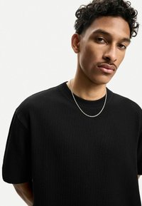 Črna pletena majica s kratkimi rokavi z teksturiranim vzorcem, okroglo izrezana, jo drži oseba, ki nosi srebrno verižico.