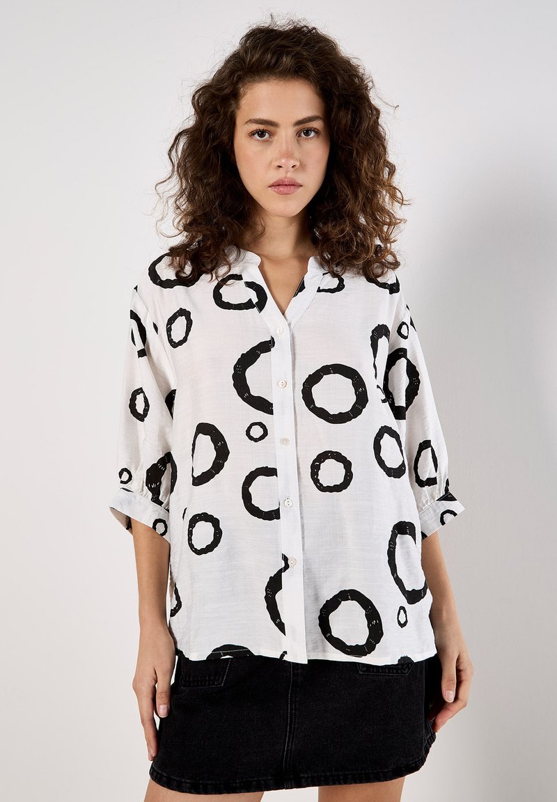 White button-up shirt featuring large black circular patterns, short sleeves, and a relaxed fit, paired with a black denim skirt.