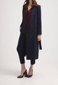 Long navy coat over a burgundy top, paired with navy trousers. Black patent leather high-heeled shoes, with a modern, tailored design.