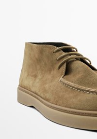 Brune ruskindsankelstøvler med rund tå, to snørehuller og kontrasterende syninger langs sålen. Glat tekstur og minimalistisk design.