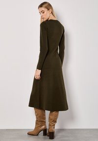 Langärmliges, olivgrünes Strickkleid mit figurbetontem Oberteil und ausgestelltem Rock; getragen mit hohen, beigen Stiefeln mit Absatz. Glatte Textur.