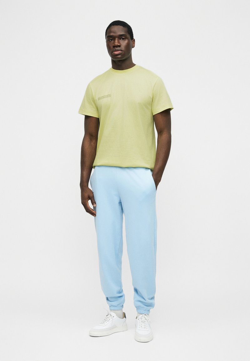 Άνδρας που στέκεται φορώντας ένα ανοιχτό πράσινο κοντομάνικο μπλουζάκι, ανοιχτό μπλε παντελόνι τζόγκινγκ και λευκά αθλητικά παπούτσια μπροστά σε ένα απλό φόντο.