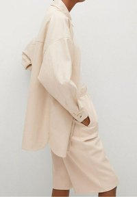 Osoba ubrana w luźną, beżową koszulę z długim rękawem oraz dopasowane szorty do kolan, z rękami w kieszeniach, stojąca na tle jasnego tła.