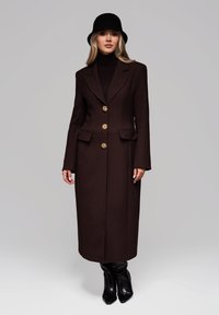 Brun uldblandet frakke med to for- lommer, gyldne knapper og en skræddersyet design. Bæres med en sort turtleneck og hat.