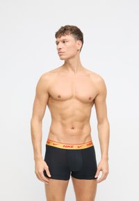 Čierne boxerky s červeným a zlatým pásom s logom Nike. Model má vypracovanú postavu a stojí v profile proti jednoduchému pozadiu.