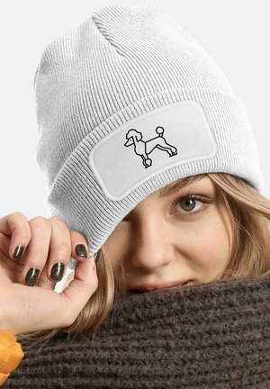 White knit beanie with a textured design, featuring a patch with a black outline of a poodle. A person holds the beanie up to their face.