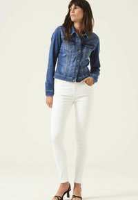 Donkerblauwe denimjack met knoopsluiting, twee borstzakken en contrasterende stiksels; gecombineerd met witte skinny jeans en zwarte sandalen.