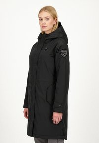 Sort vandtæt parka med hætte, der har en sidelomme med lynlås, knaplukning og logo-patch på venstre ærme. Glat stoftekstur.