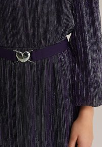 Violeta josta ar sirdi formas sudraba sprādzi, kombinēta ar faktūru melnu kleitu, kurai ir vertikālas metāliskas diegu svītras un pūkainas piedurknes.