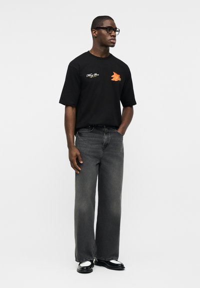 Άνδρας με μαύρο μπλουζάκι με σχέδιο, φθαρμένο γκρι τζιν φαρδιάς γραμμής, μαύρα και λευκά παπούτσια και γυαλιά, στέκεται με το ένα χέρι στην τσέπη.