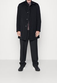 Schwarzer Mantel mit Knopfschluss und hohem Kragen, getragen über einem gemusterten Pullover und schwarzen Hosen. Fußbekleidung: schwarz-weiße Sportschuhe.