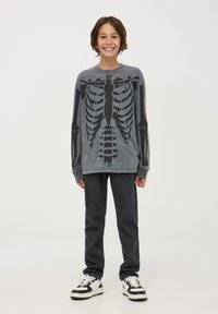 Szara koszula z długim rękawem z czarnym nadrukiem szkieletu, w zestawie z ciemnymi dżinsami i czarno-białymi trampkami. Gładka tekstura, luźny krój.