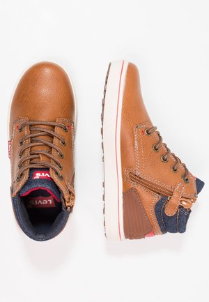 Bruna läder hög-top sneakers med vita sulor, med snörningsdesign, denimdetaljer och en dekorativ dragkedja på sidan.