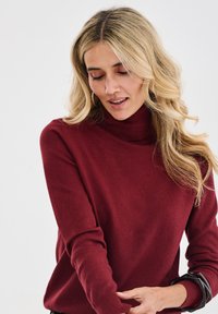 Frau mit langen, welligen blonden Haaren, die einen burgunderfarbenen Rollkragenpullover trägt und ihren Ärmel vor einem schlichten weißen Hintergrund anpasst.