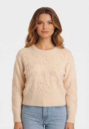 Vrouw draagt een beige trui met verhoogde bloemenborduursels en een lichtblauwe spijkerbroek, staand tegen een effen witte achtergrond.