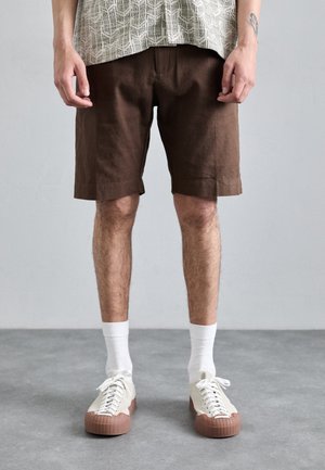 Braune Leinen-Shorts mit lockerer Passform, ausgestattet mit seitlichen Taschen. Weiße Socken und cremefarbene Sneakers mit einer braunen Gummisohle runden das Outfit ab.