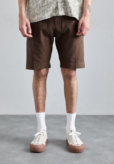 Shorts en lin marron à coupe décontractée, dotés de poches latérales. Des chaussettes blanches et des baskets crème avec une semelle en caoutchouc marron complètent la tenue.