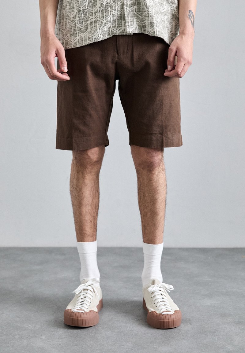 Bruine linnen shorts met een ontspannen pasvorm, voorzien van zijzakken. Witte sokken en crèmewitte sneakers met een bruine rubberen zool maken de outfit compleet.